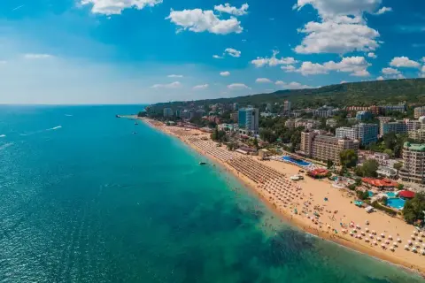 Słoneczna plaża w Złote Piaski, z mnóstwem leżaków i turystów. W tle widać hotele i zielone wzgórza. Idealne miejsce na wakacje, niedaleko lotnisko.