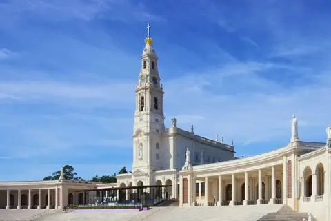 Biała bazylika z wieżą i złotą koroną na szczycie, otoczona kolumnadą. To Sanktuarium Matki Bożej Różańcowej w Fatimie, miejsce pielgrzymek.