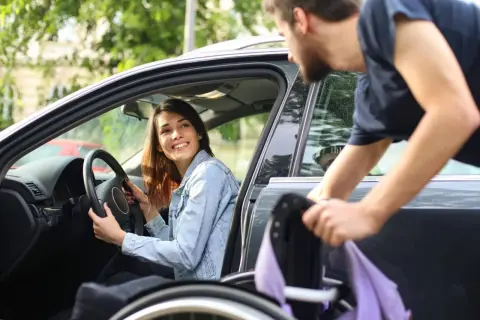 Una donna disabile sorride al volante mentre un uomo aiuta a caricare la sua sedia a rotelle in auto.