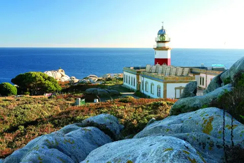 Faro en las islas Galicia, con el mar azul de fondo y rocas cubiertas de vegetación.