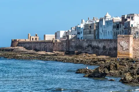 Vue des remparts d'Essaouira, avec des maisons blanches en arrière-plan. Les rochers et la mer invitent à toutes les activités.
