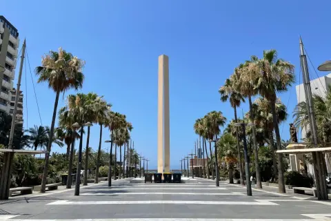 Un obelisco se alza en la plaza de las velas Almería, flanqueado por palmeras y un cielo azul despejado.