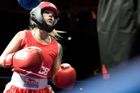 A determined female boxer in red gear and headguard prepares for a white collar boxing match, her red gloves raised.