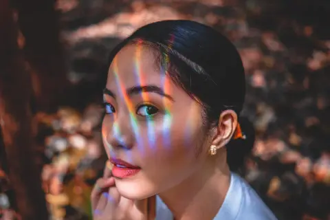 Un occhio a mandorla cattura la luce arcobaleno, riflessi colorati danzano sul suo viso mentre guarda pensierosa.