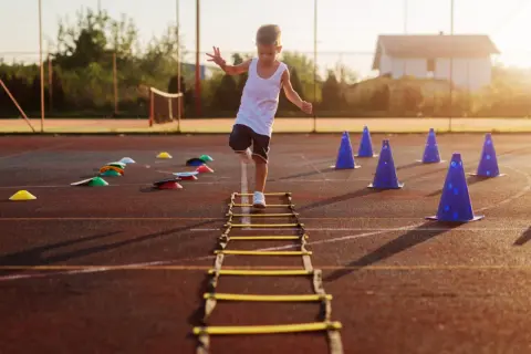 A young boy practices gross motor activities for 5 year olds, stepping through an agility ladder on a sports field with cones.