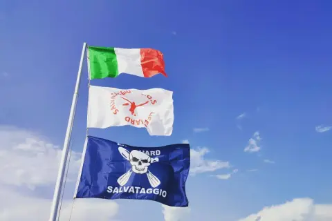 Bandiera azzurra con croce bianca, simbolo di salvataggio, sventola accanto alla bandiera italiana sotto un cielo azzurro.