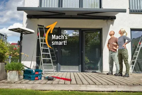 Sonnenschutz Terrasse Ideen: Mann und Frau blicken nach oben, während Werkzeug und Leiter auf der Holzterrasse bereitstehen.