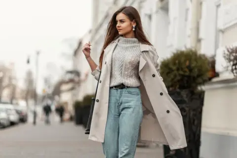 Styling Mom Jeans: Ein lässiger Look mit Strickpullover, Trenchcoat und Perlenschmuck auf der Straße.