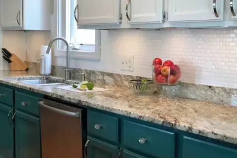 A kitchen with a white subway tile backsplash, granite countertops, and teal cabinets. A bowl of apples sits on the counter.