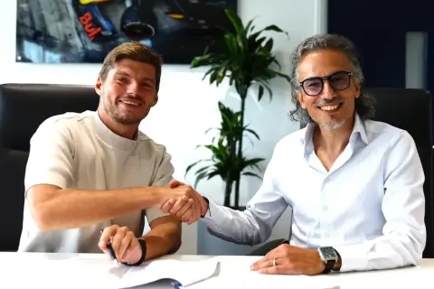 Max Verstappen sonríe mientras estrecha la mano de un hombre con gafas, sellando un acuerdo.