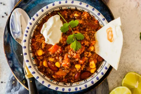 Herzhaftes Chili sin Carne mit Quinoa, Bohnen und Mais, garniert mit Sauerrahm und Koriander. Dazu eine Tortilla-Ecke und Limettenspalten.