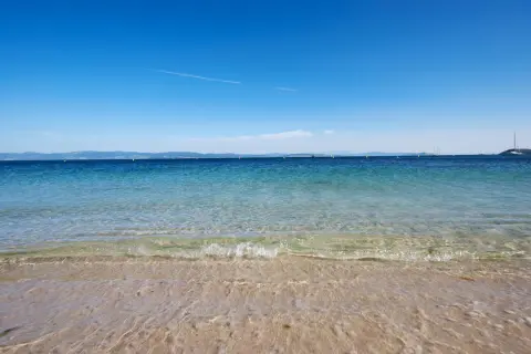 Aguas cristalinas en la playa milide, con veleros navegando en el horizonte bajo un cielo azul intenso.