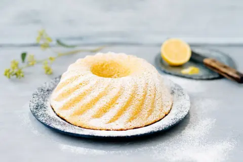 En vackert pudrad sockerkaka med citron, serverad på en mörk tallrik med en citronskiva och en kniv i bakgrunden.