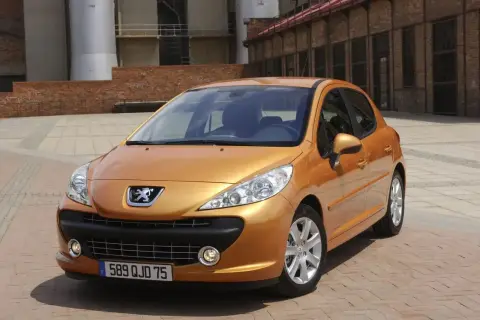 Pomarańczowy Peugeot 207 1.4 benzyna zaparkowany na placu z ceglaną posadzką, w tle budynki przemysłowe.