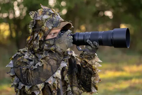 Osoba w kamuflażu z liśćmi czeka na ptaki, gotowa do fotografowania.