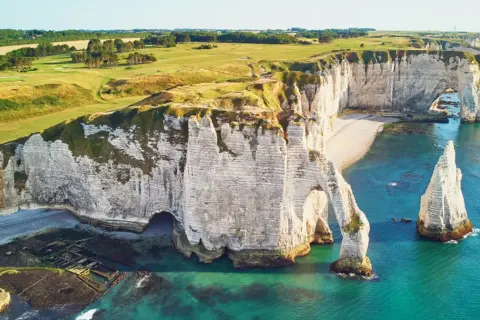 Wapienne klify nad kanałem La Manche, z łukiem skalnym i iglicą. Na szczycie klifów zielone pola golfowe.