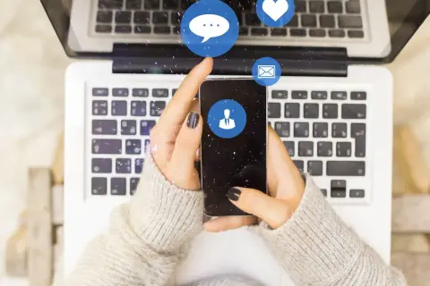 Dłonie trzymające smartfon nad klawiaturą laptopa. Ikony czatu, serca i maila unoszą się w powietrzu. Czy jak usunę czat na Messengerze to druga osoba to widzi?