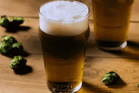 Bicchiere di birra dorata con schiuma bianca, circondato da luppoli verdi su un tavolo di legno. Perfetta per una dual IPA.