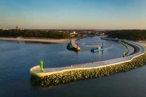 Port w Mrzeżynie z falochronami i latarniami morskimi. Widoczna łódź pogłębiarka. To jedne z mrzeżyno atrakcje.