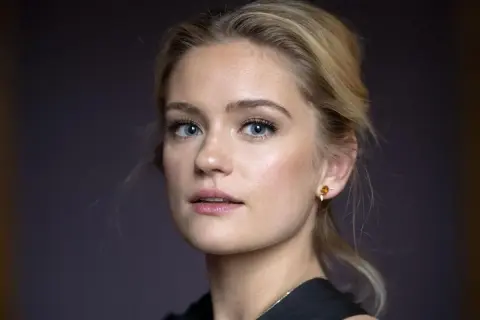Alicia Agneson, med blå ögon och blont hår uppsatt i en hästsvans, bär en guldig örhänge.