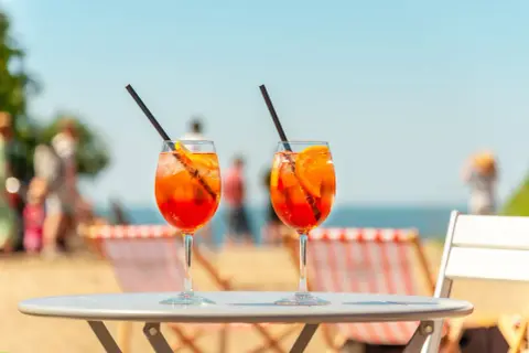 Dwa kieliszki Aperol Spritz z pomarańczą i słomką na stole, w tle plaża i morze.