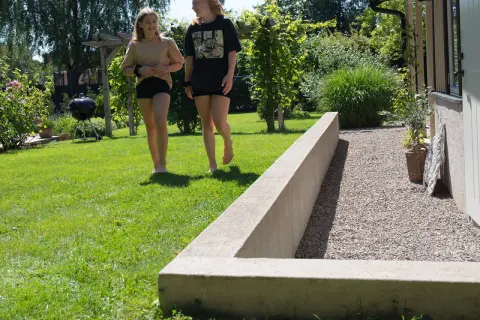 Två flickor går på en gräsmatta bredvid en nybyggd mur i en solig trädgård.