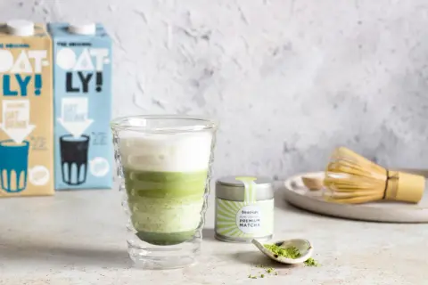 Warstwowy matcha latte w szklance z podwójnymi ściankami, obok puszka z matchą i bambusowa miotełka.