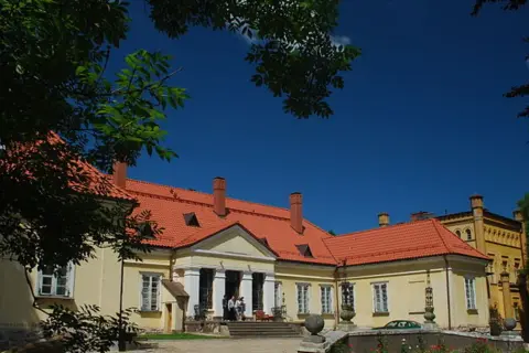 Pałac w Waplewie z czerwonym dachem i żółtymi ścianami, otoczony zielenią, pod błękitnym niebem.