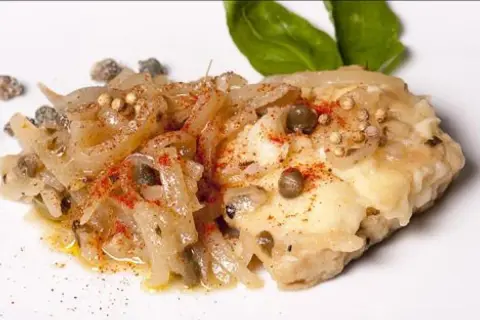 Un morceau de lotte poisson, cuisiné avec des oignons, des câpres et des épices, garni de feuilles de basilic.