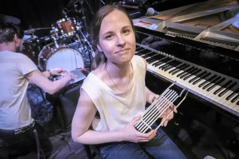 Hania Rani, przy fortepianie, trzymając w dłoniach nietypowy instrument, gotowa do tworzenia swoich unikalnych utworów.