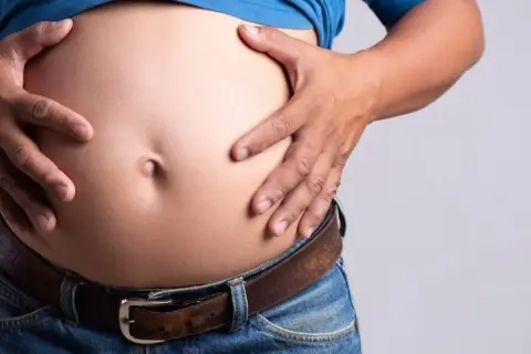 Un homme, je suis mince avec un gros ventre, tient son abdomen proéminent au-dessus de son jean et de sa ceinture.