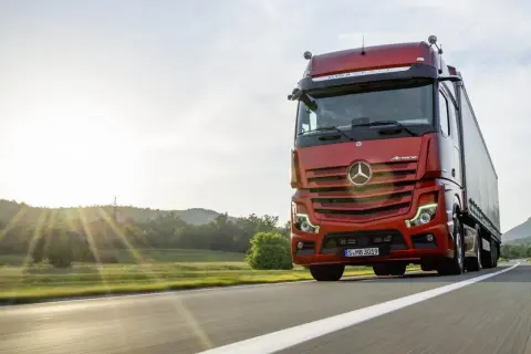 Un camion rouge vif roule sur une route ensoleillée, le soleil couchant illuminant le paysage. Ce camion sans rétroviseur semble prêt pour une longue route.