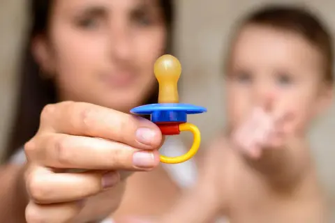 A mother holds a colorful pacifier, perhaps contemplating extended pacifier use for her baby, who is blurred in the background.
