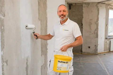 Maler grundiert mit einer Rolle die Wand. Er hält einen Eimer mit "StoPrim GT" in der Hand.