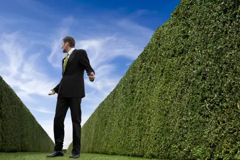 Mann im Anzug steht vor einer Hecke, die wie ein Labyrinth wirkt. Er wirkt ratlos, als ob er eine christliche Sekten Liste sucht.