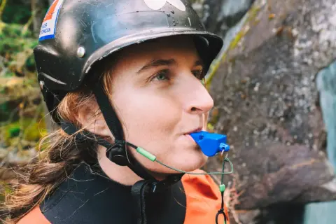 Persona con casco y traje naranja sopla un silbato de emergencia azul, listo para cualquier aventura acuática.
