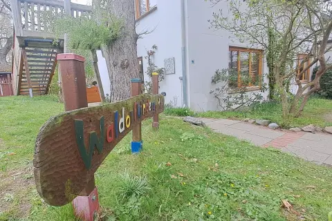 Holzschild "Waldorfkindergarten" vor einem Gebäude mit Spielturm. Mögliche Nachteile: wenig Struktur, viel Freiheit.