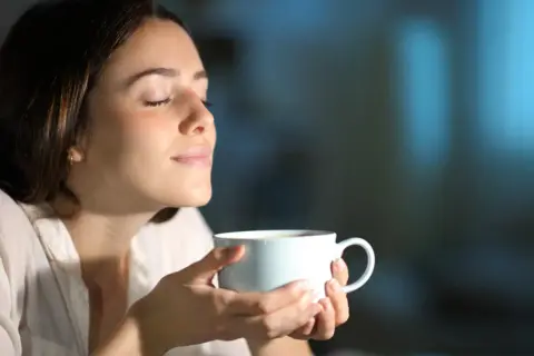 Kobieta z zamkniętymi oczami wdycha aromat napoju. Może to być kawa lub napar z ziół na pobudzenie.