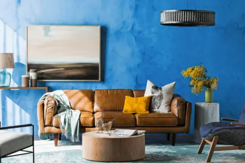 Ein Wohnzimmer mit leuchtend blauer Wand. Ein braunes Ledersofa, ein gelbes Kissen und ein Bild mit Landschaftsmotiv. Welche Farbe passt gut zu Blau? Hier: Gelb und Braun.