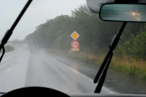 Conduire sous la pluie sur une route mouillée, avec des essuie-glaces en action et un panneau de rappel de vitesse 110.