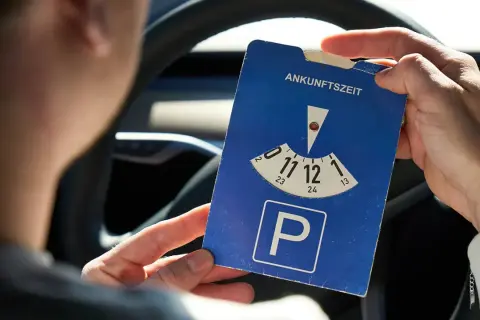 Person hält eine Parkscheibe mit der Aufschrift "Ankunftszeit" und dem Parkplatzsymbol.