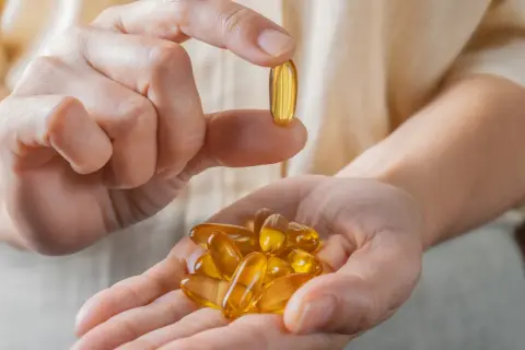 Dłoń trzyma kapsułkę z omega-3, zarejestrowaną jako lek. W drugiej dłoni garść takich tabletek.