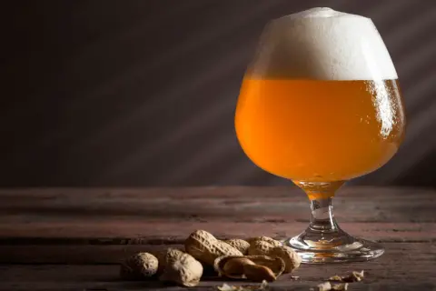 Un bicchiere di keller beer dorata con schiuma bianca, accompagnata da arachidi su un tavolo di legno scuro.
