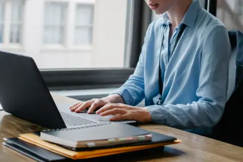Osoba w niebieskiej koszuli pracuje przy laptopie, tworząc arkusz xlsx. Na biurku leżą notatniki i teczka.