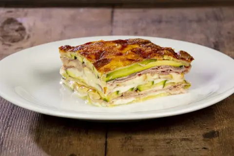 Una fetta di parmigiana di zucchine bianca alla napoletana, stratificata con prosciutto e formaggio fuso, su un piatto bianco.