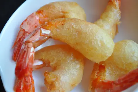 Plusieurs beignets de crevette dorés et croustillants, avec des queues de crevettes roses qui dépassent, servis dans une assiette blanche.
