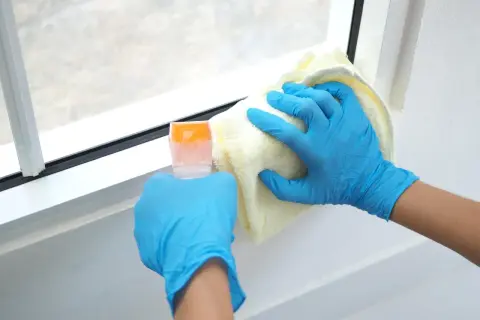 Ręce w niebieskich rękawiczkach czyszczą białe ramy okienne plastikowe. Użyj delikatnego środka do czyszczenia, by przywrócić im blask.