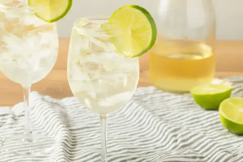 Orzeźwiający szprycer drink z lodem i plasterkiem limonki, obok butelka białego wina i połówki limonki.