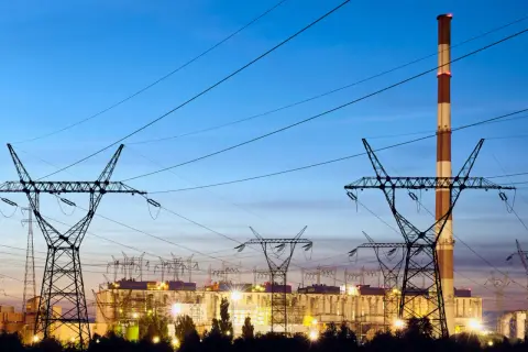 Elektrownia z kominem i liniami wysokiego napięcia. W tle widać budynki zakładu, a nad nimi niebo o zmierzchu. To obraz, który może kojarzyć się z opłatą kogeneracyjną.
