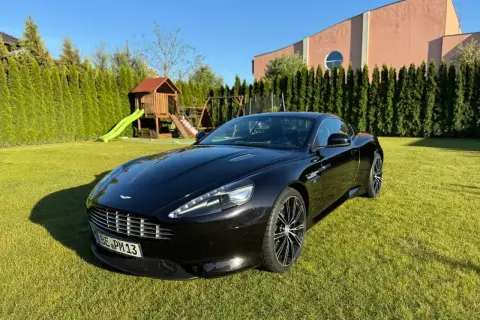 Czarny Aston Martin DBS lśni na trawniku przed domem. W tle plac zabaw i wysokie tuje.